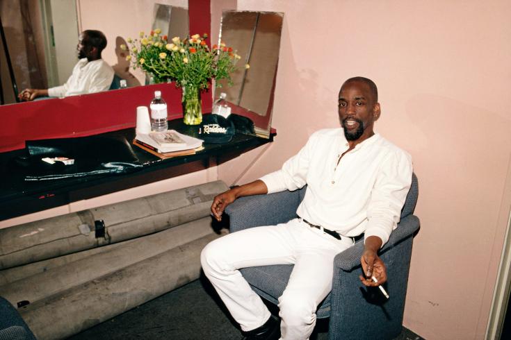 Man wearing an all white outfit sitting in a chair holding a cigarette.