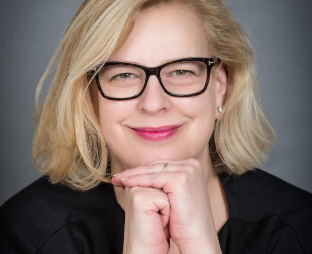 Julie Garcia, a blonde woman with glasses, smiles at the camera with her hands folded under her chin.